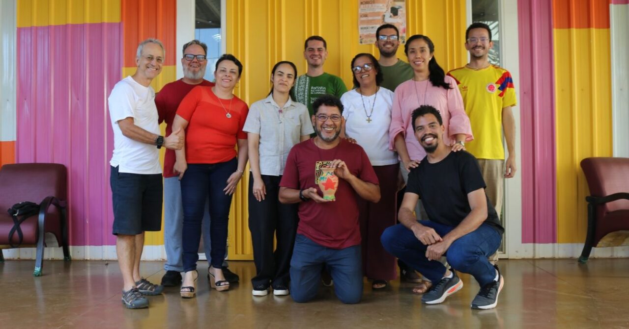 Grupo de dez pessoas posa sorridente para uma foto em frente a uma fachada colorida, com tons de amarelo, rosa e laranja. Duas pessoas estão agachadas à frente, e uma delas segura um pequeno objeto decorativo.
