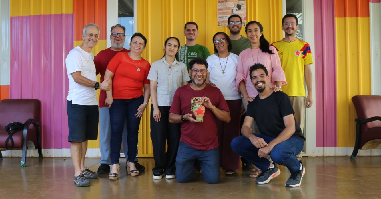 Grupo de dez pessoas posa sorridente para uma foto em frente a uma fachada colorida, com tons de amarelo, rosa e laranja. Duas pessoas estão agachadas à frente, e uma delas segura um pequeno objeto decorativo.