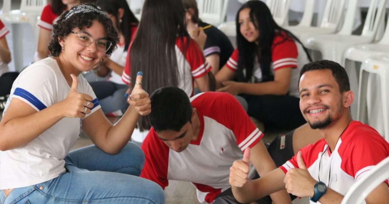 Jovens estudantes reunidos em atividade de convivência e formação, com dois participantes sorrindo para a câmera e fazendo sinal positivo com as mãos, enquanto outros conversam ao fundo.