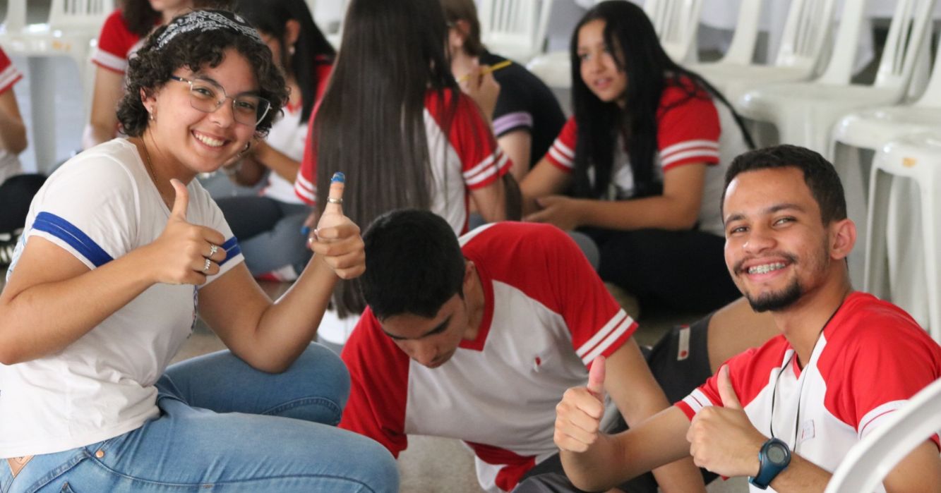 Jovens estudantes reunidos em atividade de convivência e formação, com dois participantes sorrindo para a câmera e fazendo sinal positivo com as mãos, enquanto outros conversam ao fundo.